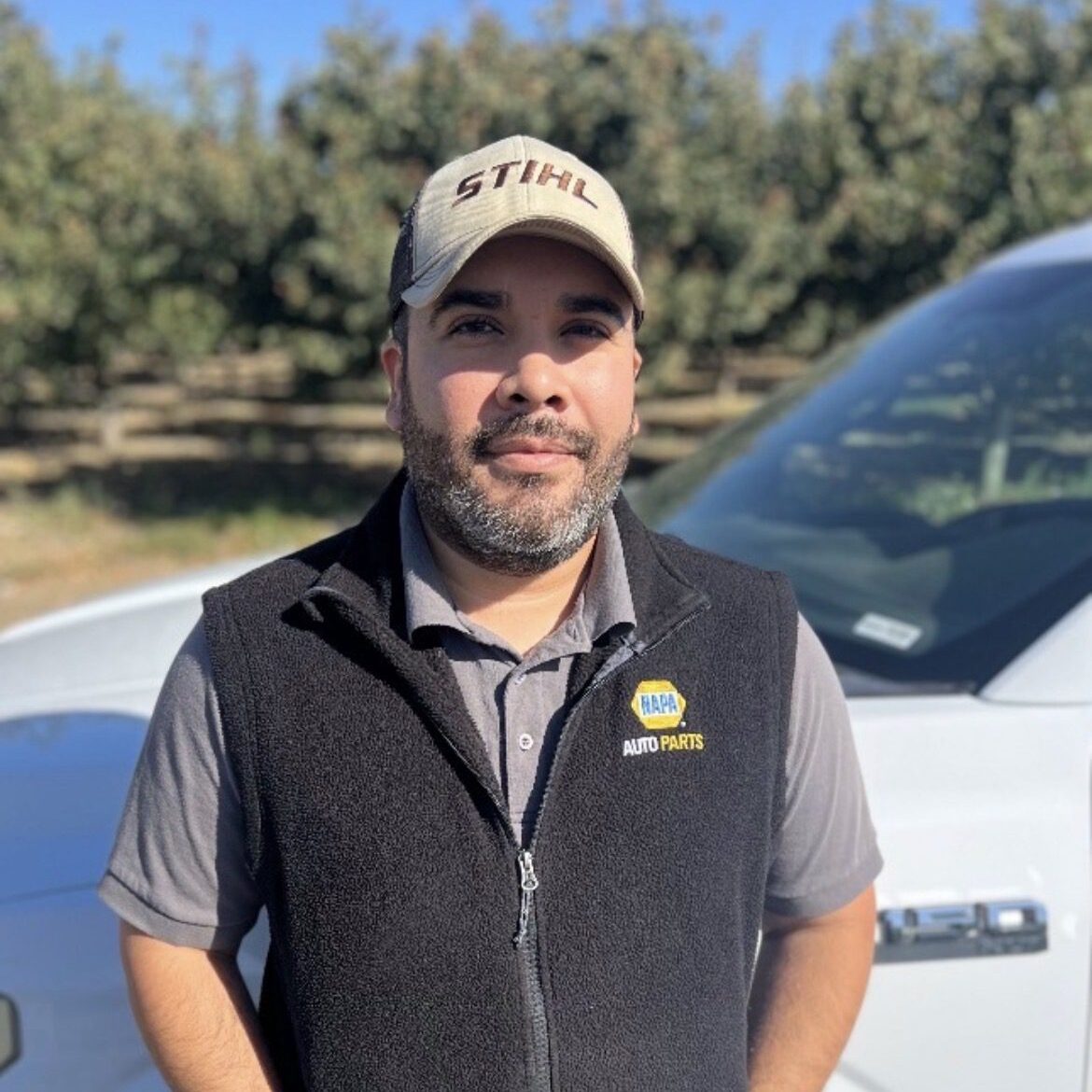 Man in a cap and vest standing outdoors near a white vehicle.